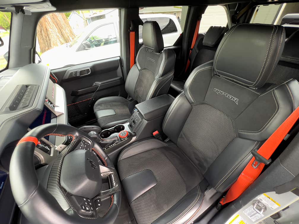 A close-up of the interior of a Ford Bronco Raptor front seats with a steering wheel in the center of the vehicle and the center console in the center of the vehicle