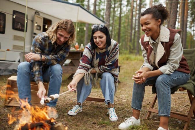 3 adults sit around a campfire with an RV in the background