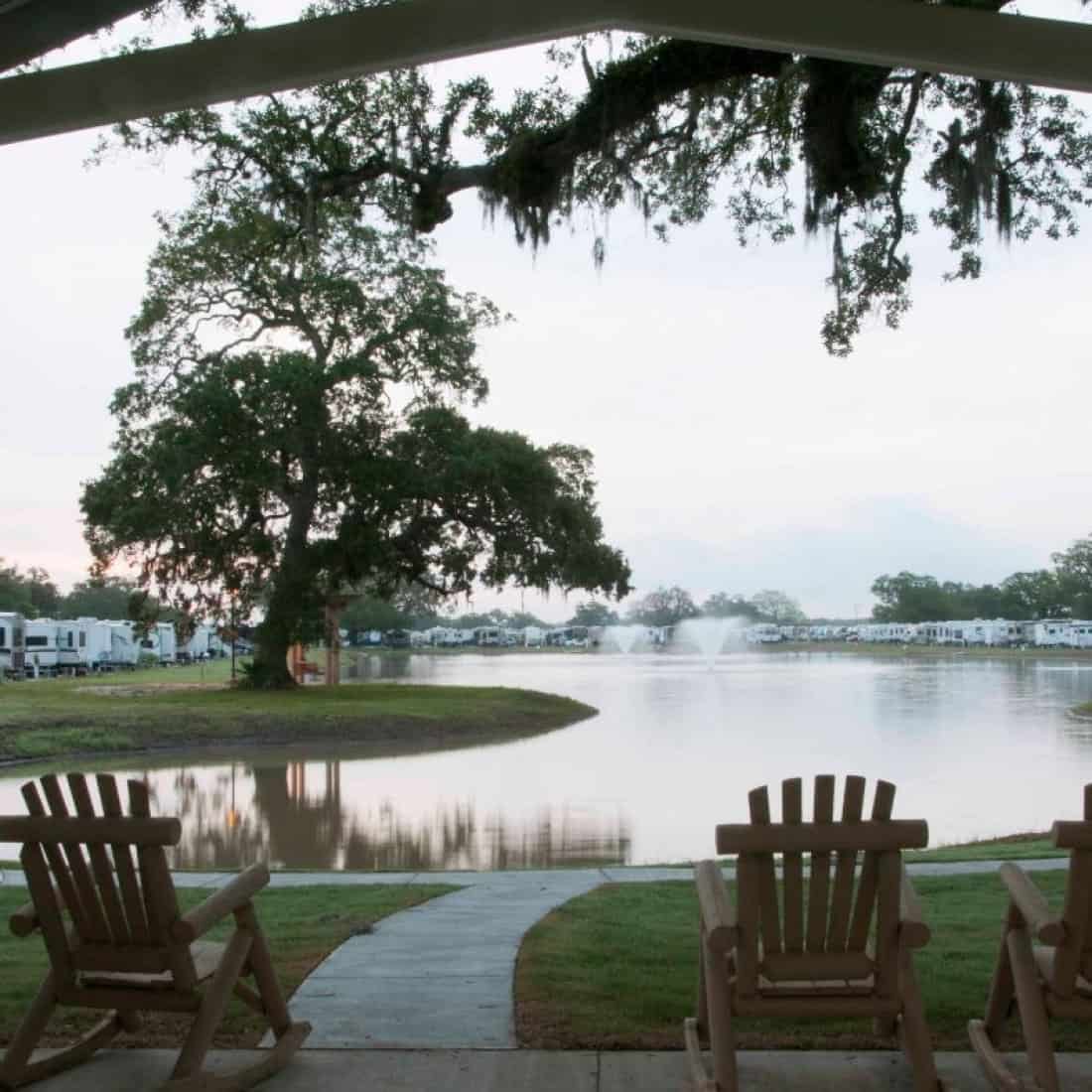 Rocking chairs facing a serene lake with fountains at Brazoria Lakes RV Resort, flanked by RVs and lush trees.