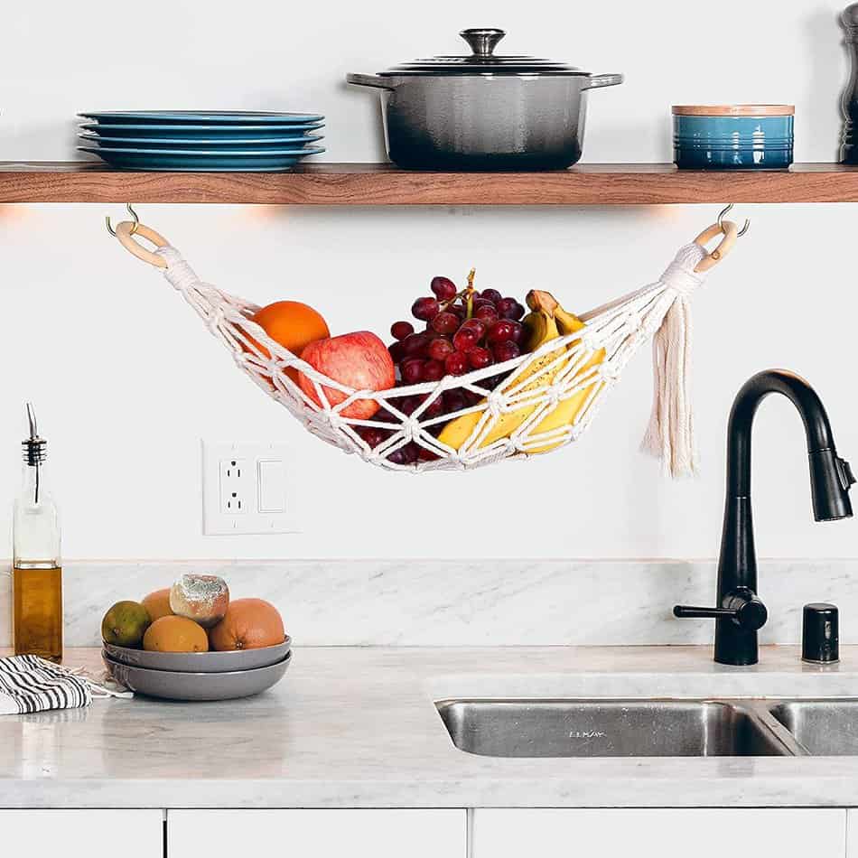 Macrame Fruit hammock with bananas and apples in it hanging under a cabinet above a kitchen sink 