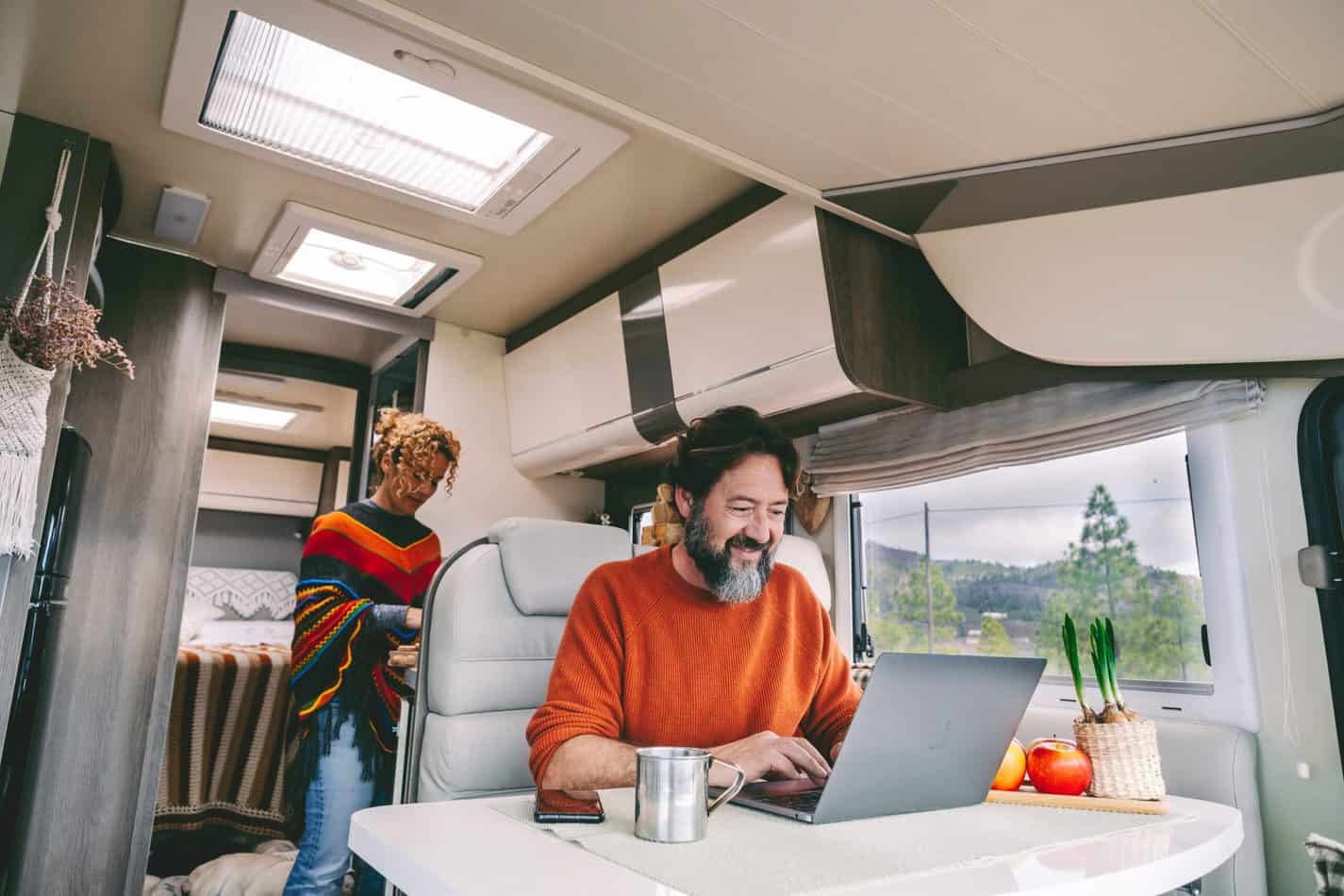 Man sitting an an RV table with a laptop, a woman standing behind him at a kitchen counter