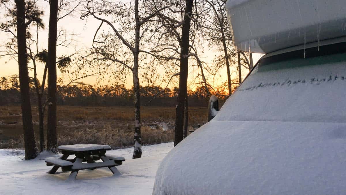 Snow covered RV camper at sunrise parked for some winter RV camping at a campground with a picnic table in the background.