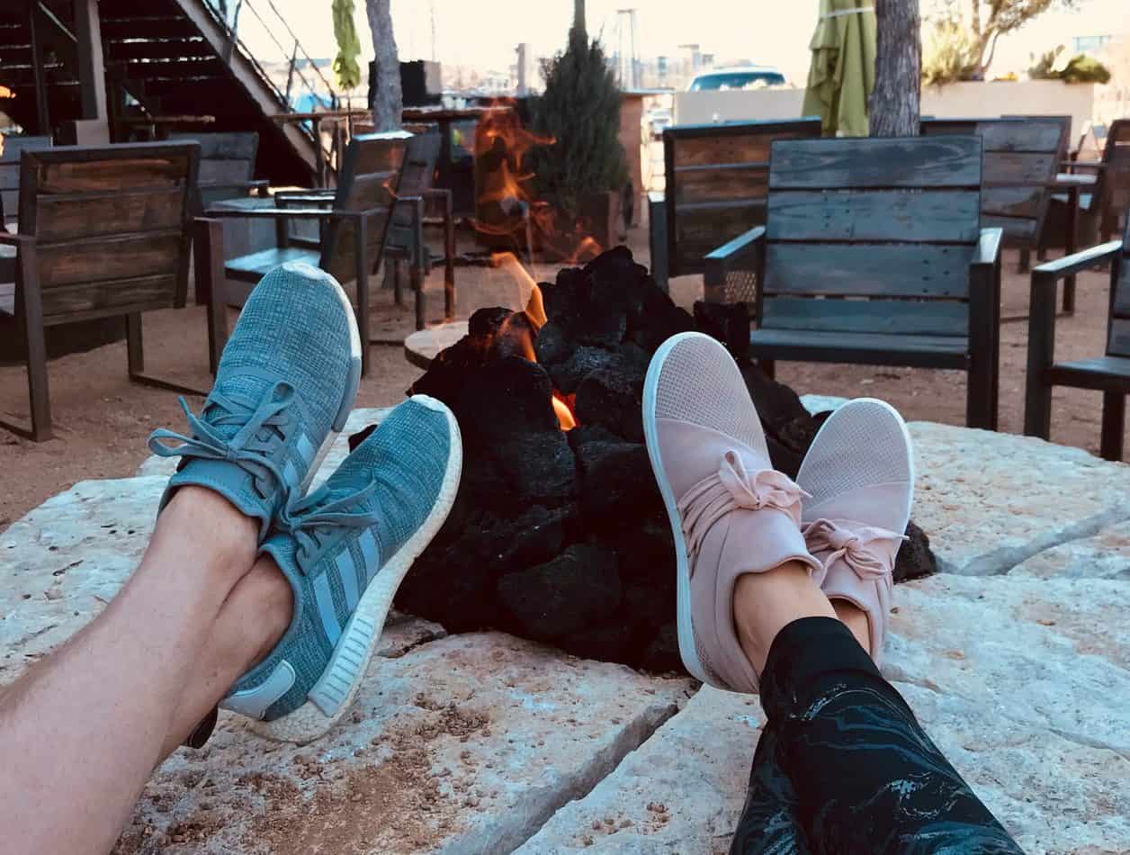 Two pairs of legs with sneakers on propped up on a fireplace with a fire